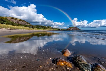Rainbow arching over a sandy beach with the ocean beyond, clear skies and gentle waves