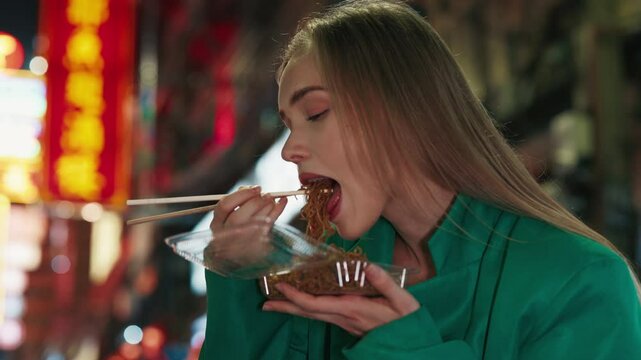 Portrait of beautiful young woman eating instant noodle with chopsticks resting in night city. Pretty female enjoys street food with great pleasure in evening urban district
