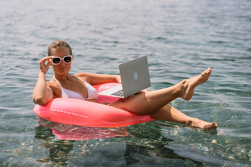 A woman is floating in a pink inflatable raft with a laptop in her hand. She is smiling and she is...