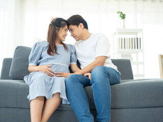 A happy Asian couple. Handsome husband and his beautiful wife pregnant sitting on sofa.