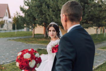 A bride and groom are walking together in a park, with the bride holding a bouquet of red flowers. The scene is romantic and happy, as the couple is getting ready to celebrate their wedding