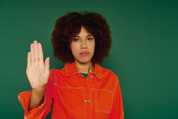 This young African American woman with curly hair showing stop gesture while dressed casually on a green backdrop.