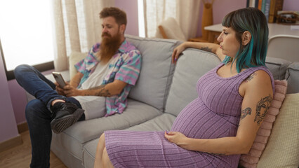 Obraz premium Pregnant woman with tattoos sitting on a couch in a living room while a bearded man next to her looks at his phone, showcasing a contemporary family moment indoors.