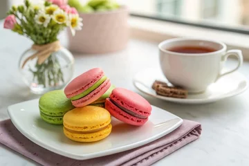 A plate holds vibrant macarons near a cup of tea and a floral vase on a table.