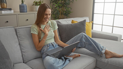 Young attractive blonde woman using a laptop in the living room of a home sitting on a comfortable couch indoors.