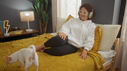 Middle-aged woman with headphones sitting on a bed in a cozy bedroom while her chihuahua dog stands...