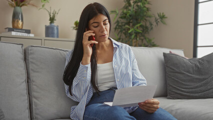 Attractive hispanic brunette woman sitting in a living room on a couch, talking on a phone while looking at a document.