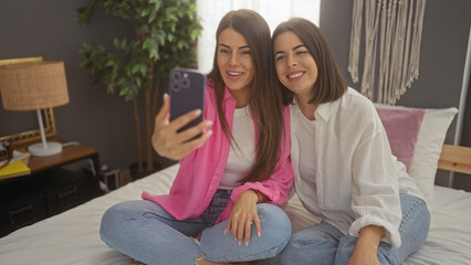 Two smiling hispanic women sitting on a bed in a cozy bedroom, taking a selfie together, capturing a moment of friendship and joy.