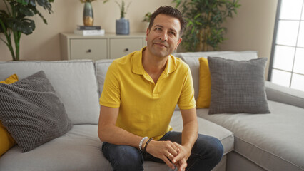 Fototapeta premium Hispanic man sitting in a modern living room, wearing a yellow shirt and jeans, with plants and cushions around, exuding a relaxed and thoughtful demeanor in a well-decorated home interior.