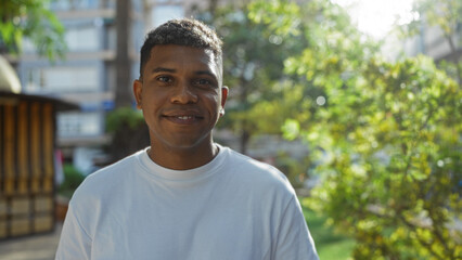 Young hispanic man smiling in an urban park with trees and buildings in the background during a sunny day