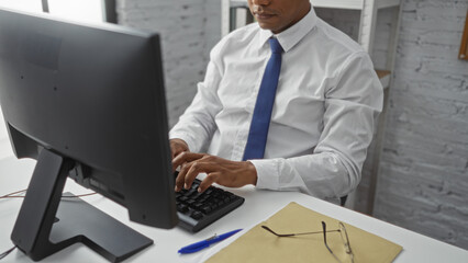 Young hispanic man working on a computer in an office setting with a file, pen, and glasses on the desk