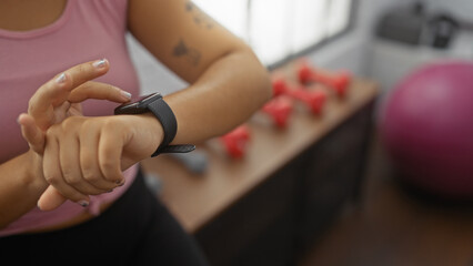 A young hispanic woman at the gym looks at her smartwatch in an indoor gymnasium, with dumbbells and exercise equipment in the background.