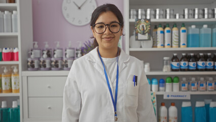 A young hispanic brunette woman in a white coat stands in a pharmacy with shelves of products behind her.