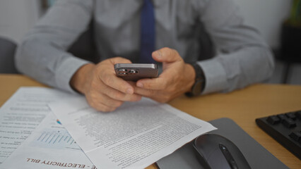 Hispanic man using smartphone in an office with documents and keyboard visible on the desk, focusing on his hands as he works indoors.