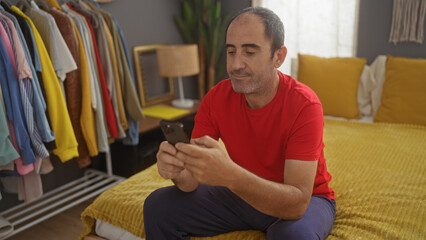 A middle-aged hispanic man sits on a bed in a cozy bedroom, looking at his smartphone, surrounded by a colorful wardrobe and warm interior decorations.