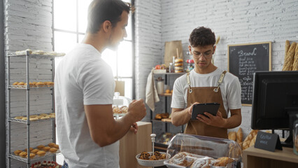 Two hispanic men in a bakery with one working as a baker wearing an apron and using a tablet while interacting with a customer indoors in a bright room surrounded by baked goods and shelves