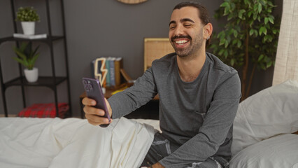 Young man taking selfie on bed in bedroom, smiling and using smartphone, surrounded by cozy home decor and indoor plants.