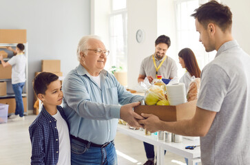 Fototapeta premium Friendly male volunteer holding box with essentials, giving free food donation to poor refugee family, smiling grandfather and grandson in charitable foundation. Humanitarian help and social support.