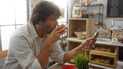 Young man drinking coffee and using smartphone in a cozy bakery with an inviting interior and delicious pastries on display.
