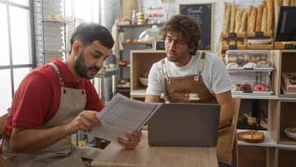 Men working together as bakers in a bakery interior, holding documents and using a laptop,...