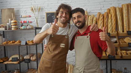 Two smiling men in aprons give thumbs up in a bakery shop interior with a display of various bread and pastries.