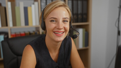 Young, blonde, woman, wearing, headset, smiling, while, working, in, office, with, bookshelves, and, documents, in, background