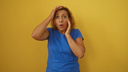 Beautiful mature hispanic woman with curly hair and a surprised expression wearing a blue shirt against an isolated yellow background.