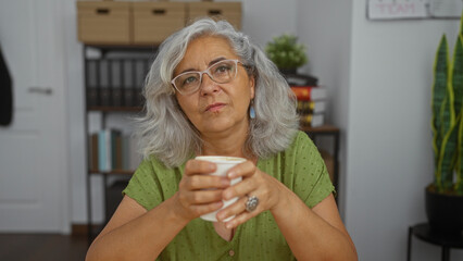 Woman with grey hair and glasses holding a cup while sitting in an office with bookshelves and a potted plant in the background.