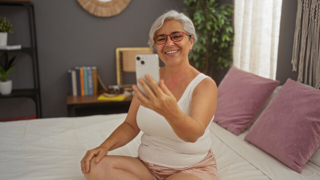 Elderly woman takes selfie with smartphone while sitting on bed in cozy bedroom at home with books on a shelf in the background