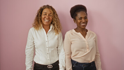 Women standing over an isolated pink background, showcasing the bond of friendship and diversity, one being african american and the other latin american, both smiling in shirts and jeans.