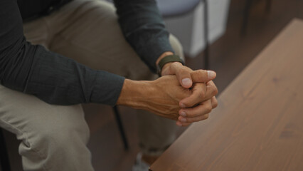 Hispanic man with clasped hands sitting in an indoor waiting room or lobby, depicting a moment of contemplation in an indoor setting.