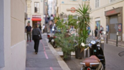 Obraz premium Blurred street in marseilles with woman walking along urban sidewalk surrounded by defocused buildings and plants in soft sunlight, creating a bokeh effect.