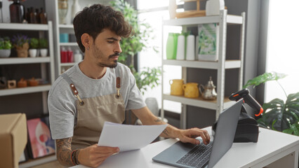 Young man with a beard holding a document while using a laptop in a well-decorated shop with shelves full of various home decor items. © Krakenimages.com