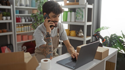 Young hispanic man with beard working on laptop and talking on phone in home decor store interior