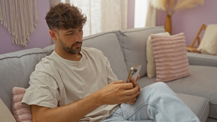 Obraz premium Young man with short curly hair sitting on a gray couch using a smartphone in a cozy living room with pink cushions and decorative elements