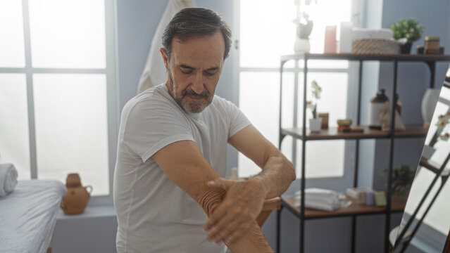 Mature hispanic man applying cream indoors at spa wellness center surrounded by tranquil decor and soft lighting emphasizing relaxation and self-care in a serene environment