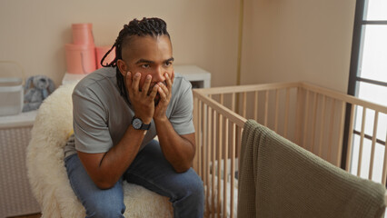 Young man with braids sitting thoughtfully in a bedroom beside a cradle, surrounded by soft lighting, conveying a sense of contemplation in a cozy indoor setting.