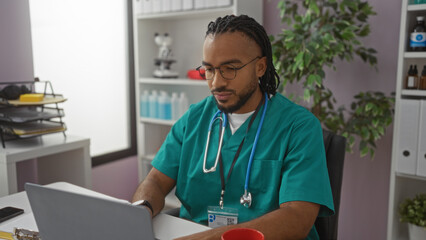 Young man with braids and glasses in a hospital clinic wearing scrubs with a stethoscope using a...