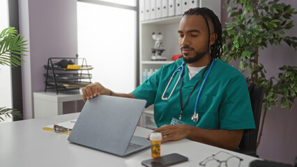 Man sitting in clinic wearing green scrubs with stethoscope while using a laptop, highlighting a...