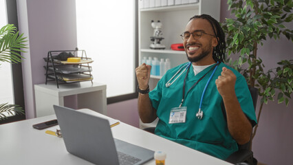 Fototapeta premium Young man in medical scrubs celebrating success in a clinic office setting with laptop and stethoscope, expressing happiness in a professional healthcare environment.