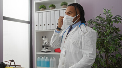 Man wearing medical mask in clinic adjusting it with focus on stethoscope suggesting professional environment indoors with organized shelves and natural light.