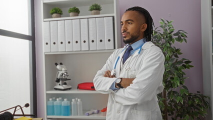 Confident man in clinic wearing lab coat stands with arms crossed beside shelves filled with...