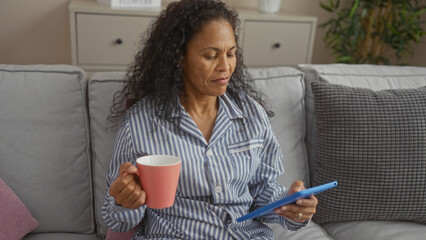 Woman relaxing in living room with coffee and tablet, wearing pajamas, middle-aged, indoors, home, hispanic, mature, african american - Powered by Adobe