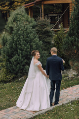 A bride and groom are walking down a brick path in front of a house. The bride is wearing a white dress and a veil, while the groom is wearing a blue suit. They are holding hands and smiling