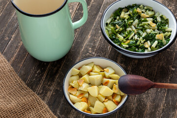 Two enameled bowls, one with boiled potatoes and fried garlic, the second with cooked leaf beet and garlic. Wooden spoon, enamel jug with water, and jute fabric. Country food.