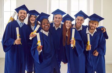 Happy diverse university friends in caps and gowns celebrating graduation together. Indoor group...