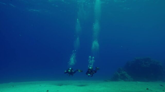 Pair of scuba divers swim over sandy bottom, approaching and waving greetings, in the blue depths sea, Slow motion of Scuba divers swim in the deep, a long trail of air bubbles rising above them