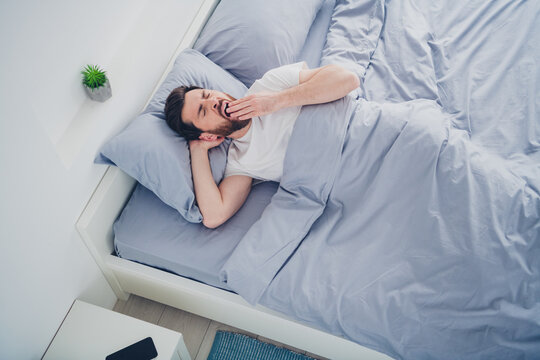 Top view photo of funny young man nightwear pajama lying bed yawning indoors house apartment room