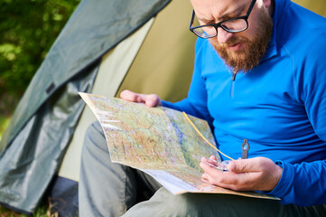 Close up man in glasses closely examines map while holding compass. He sits by his tent, deeply...