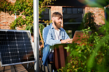 Man works on laptop at outdoor round table, with glass of iced coffee. Solar panel nearby,...